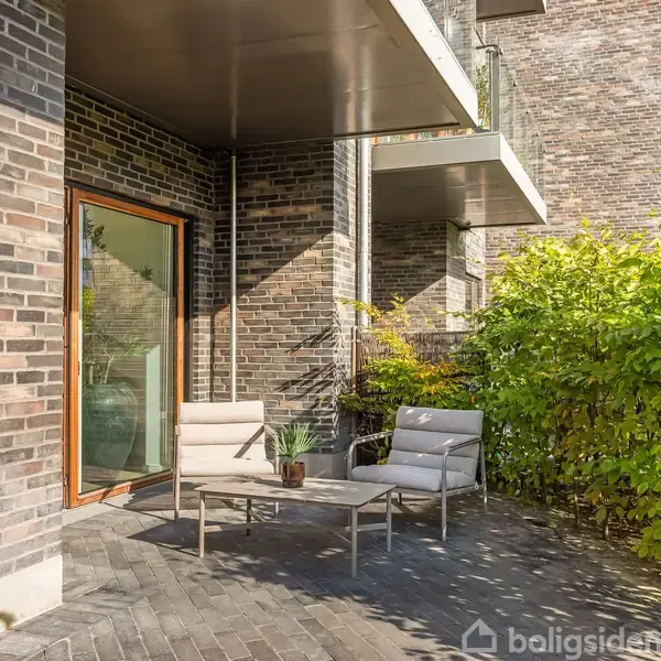 To udendørs stole står på en flisebelagt terrasse ved siden af en moderne murstensbygning. En lille bordplanter pryder bordet imellem stolene, omgivet af grønt løv og buske.