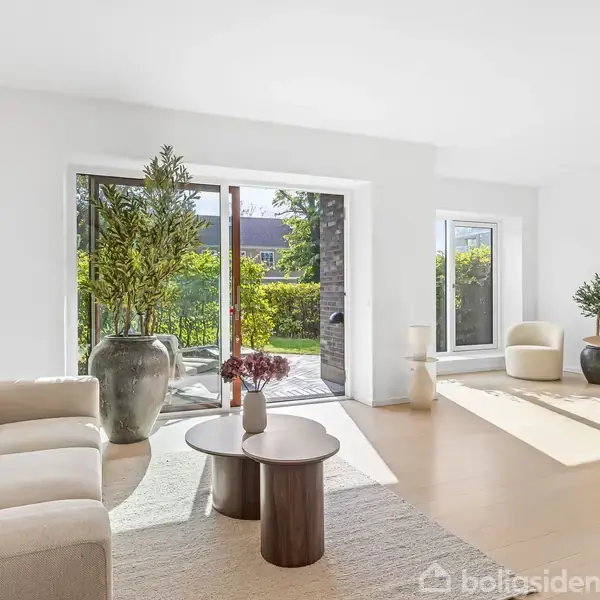 En lys stue med en beige sofa og runde borde på et tæppe. Store vinduer åbner mod en terrasse med grønne planter. Et spisebord står til højre med en vase med blomster.