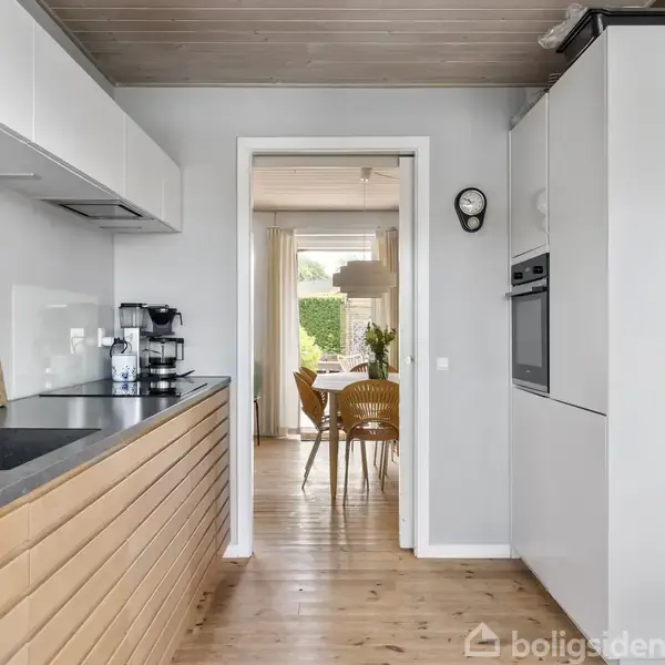 Et moderne køkken med hvide skabe og en sort bordplade. Køkkenet har en indbygget ovn, kaffemaskine og en håndvask. Gennem døren ses en spisestue med et bord og stole.