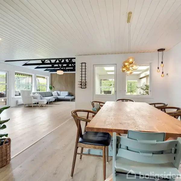 En træspisebord med forskellige stole står i et moderne, lyst rum. I baggrunden er der en åben stue med en lysegrå sofa, omgivet af store vinduer og synlige tagbjælker.