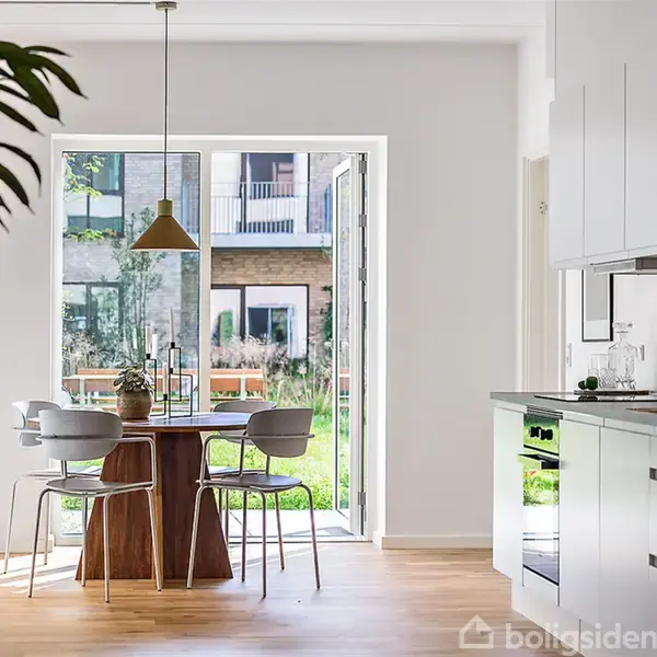 Et rundt spisebord med stole står i køkkenet nær en stor glasdør. Sollys skinner ind fra en have udenfor. Moderne indretning med trælaminatgulv og hvide køkkenskabe.