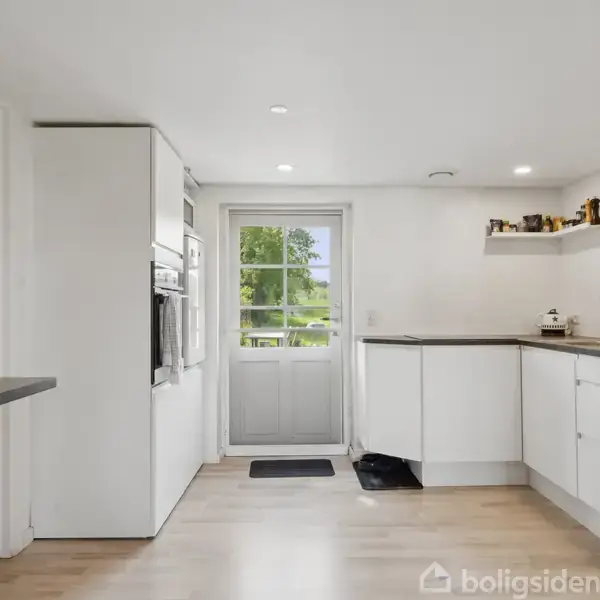 Et moderne køkken med hvide skabe og en sort bordplade, indeholder en indbygget ovn og opvaskemaskine. Døren åbner til en have, og en hylde med køkkenartikler hænger på væggen.