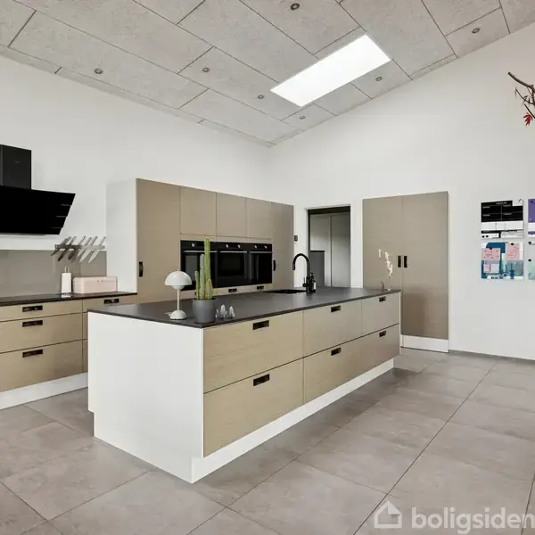 Køkkenø står i centrum, udstyret med indbygningsovne og kogeplade; opsat i et moderne rum med hvidmalede vægge, store grå fliser og naturligt lys fra ovenlysvindue. En plante ses til højre.