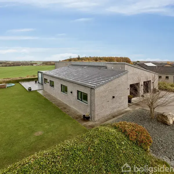 En moderne, grå murstensvilla står i stille omgivelser med velholdt græsplæne og buske. Et langt fladt tag dækker huset, med store vinduer, omkranset af åbne marker og få naboer.