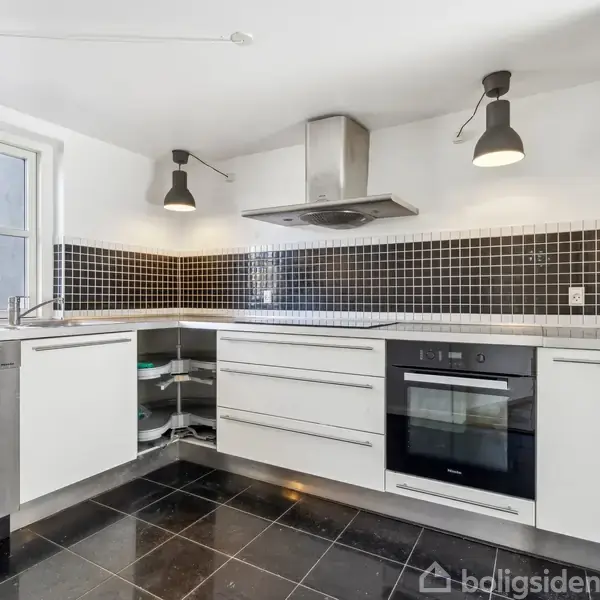 Et moderne køkken med hvide skabe og integrerede apparater. Kogeplade og ovn indbygget i bordpladen. Sorte fliser på gulvet, og væggen har et sort-mosaik flisemønster.