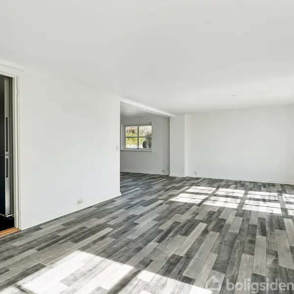 Et rummeligt, tomt værelse med gråligt trægulv, modtager sollys gennem store vinduer. Til venstre ses en åbning ind til et moderne køkken med stålelementer.