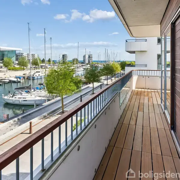 Altan med trædækning strækker sig langs en moderne bygning, udsigt mod en marina fyldt med både, kæmpe moderne bygninger i baggrunden, og klart vejr med få skyer.
