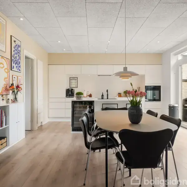 Spisebord med sorte stole står i et køkken-alrum. Bordet har en vase med blomster. Væggene er dekoreret med kunstværker. Store vinduer i baggrunden giver udsigt til terrassen.