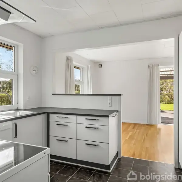 Køkken med hvide skabe, sort bordplade og stålvask, står i et rum med hvide vægge. Lys træparket og vinduer med udsigt til grøn beplantning ses i baggrunden.