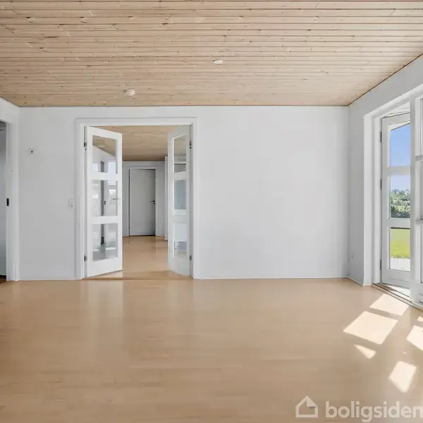 Stue med trægulv, hvor dobbeltdøre fører til et tilstødende rum. Store vinduer lader sollys strømme ind, og der er en udendørs udsigt gennem vinduerne.