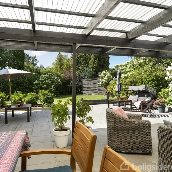 Terrasse med havemøbler står under en pergola. Møblerne omfatter stole, et bord med melon, og parasoller. Omgivet af frodige planter og blomster, skaber det en hyggelig udendørs atmosfære.