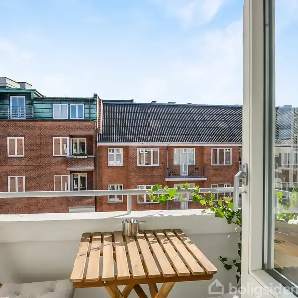 Et lille altanbord med to stole står på en balkon. Bordet er dekoreret med en lille plante. I baggrunden ses røde murstensbygninger med flere etager af lejligheder.