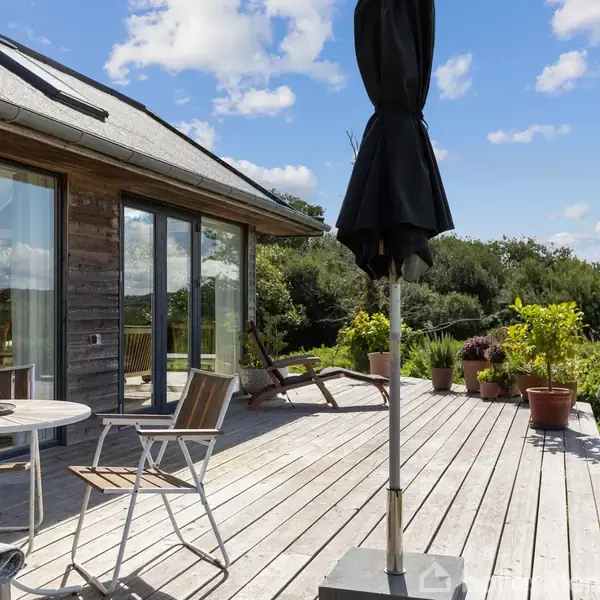 Træterrasse med havemøbler og en parasol står foran et moderne hus med store vinduer. Sollys oplyser området, der er omgivet af frodig grøn beplantning og en blå himmel.