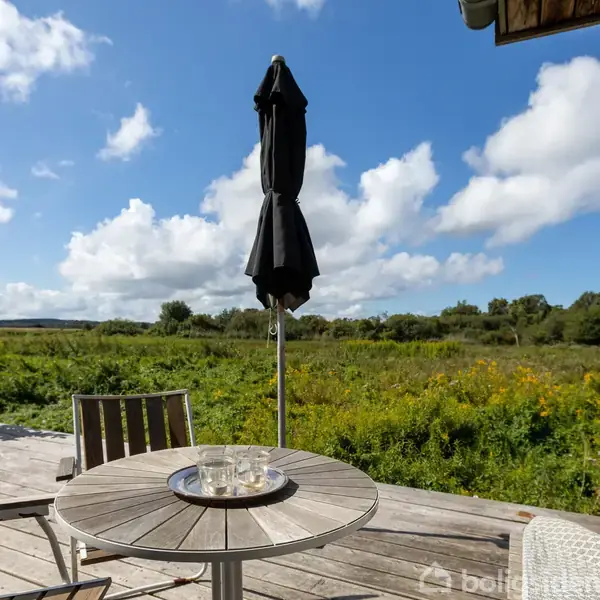 Et rundt bord med glas og bakke står på en træterrasse. En sort parasol er foldet sammen. I baggrunden ses en frodig eng under en blå himmel med skyer.