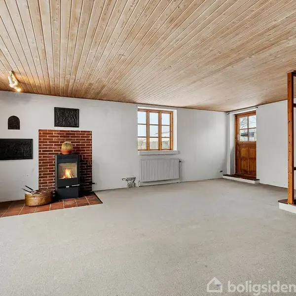 Brændeovn op ad en murstensvæg, står i en stue med trægulv og loft, store vinduespartier på venstre væg, hvide vægge, og en trædør i baggrunden.