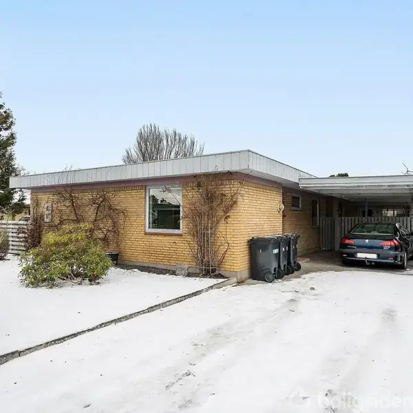 Et hus af mursten står stille omgivet af sne. En bil er parkeret i en carport ved siden af huset. Træer og buske i baggrunden.