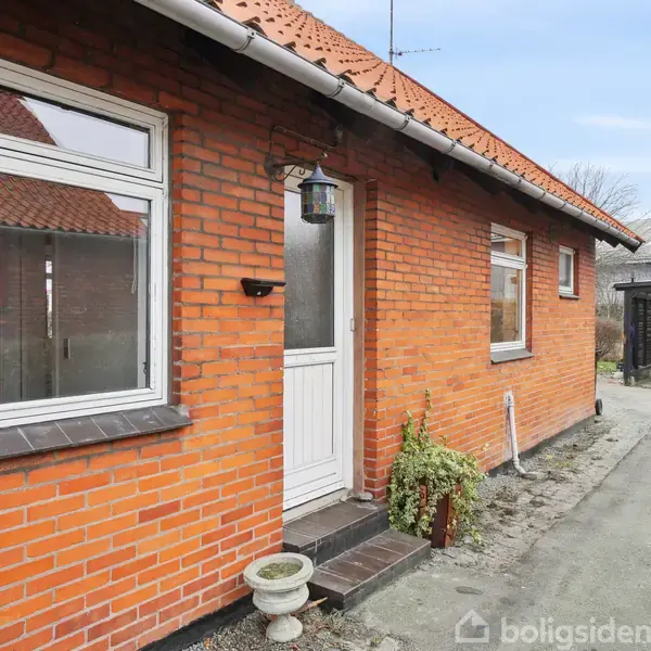 Røde murstenshus med hvid hoveddør omgivet af to vinduer. En lille blomsterpotte står ved døren. En grå indkørsel leder til en carport i en villakvarter.