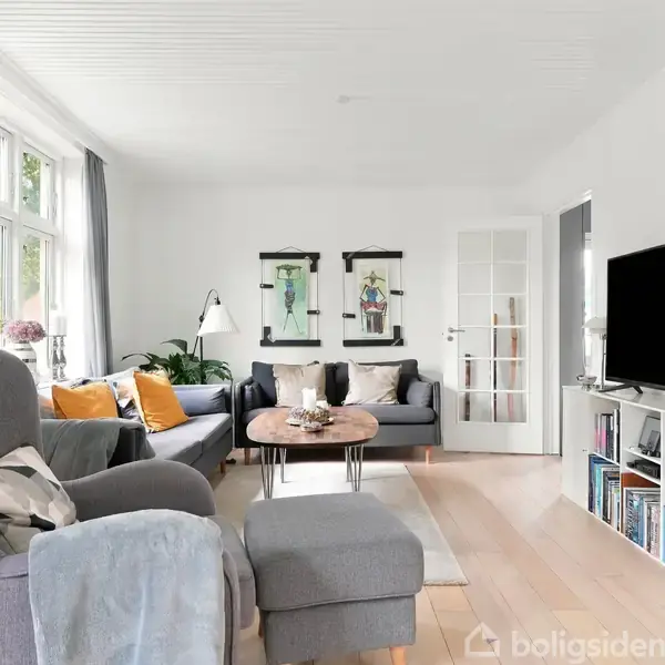 Gråt sofahjørne med puder og tæppe, imødekommende stue med store vinduer, lyse vægge, træstige, gulvlampestander, fladskærms-TV, bogreol, kunst på væggene, grøn plante.