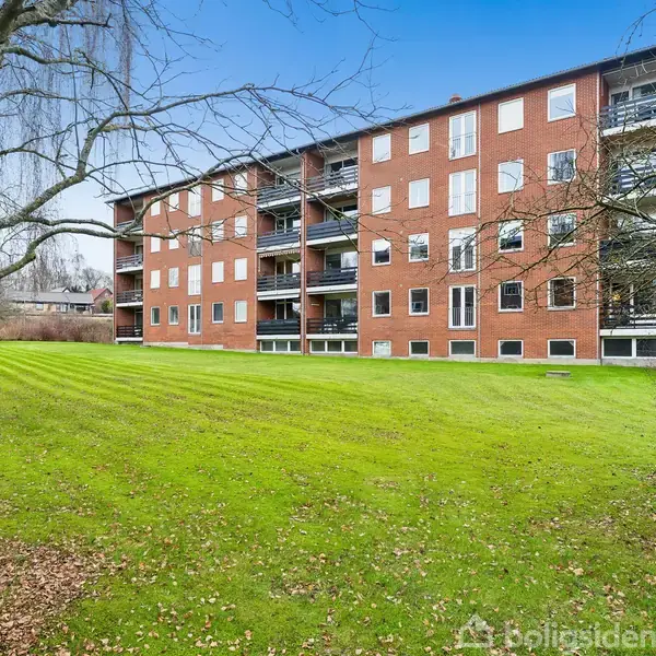 En fire-etagers murstensbygning med altaner står i forgrunden. Omkring bygningen er en grøn græsplæne med bartræer. Himlen er klar og blå.