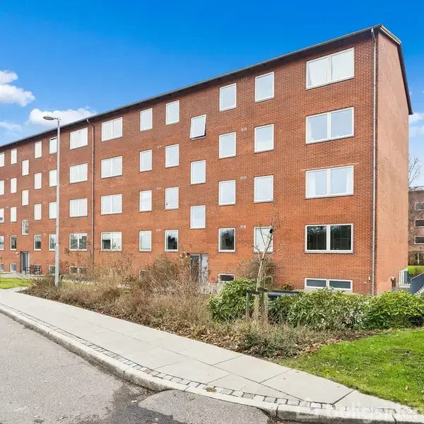 En femetagers murstensbygning står stille på en stille gade. Den er omgivet af beskeden beplantning og et åbent område under en klar blå himmel med spredte skyer.