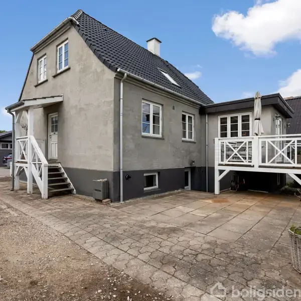 Et gråmalet hus med sort tag viser trappen op til hoveddøren. Ved siden er en terrasse med hvidt stakit. Omgivet af gårdsplads og blå himmel.