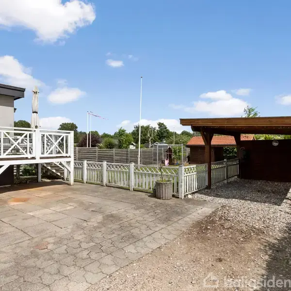 Terrasse med hvide rækværk, delvist overdækket carport til højre, beliggende i en have med grønne buske og græsarealer. Skyfri blå himmel med få skyer.