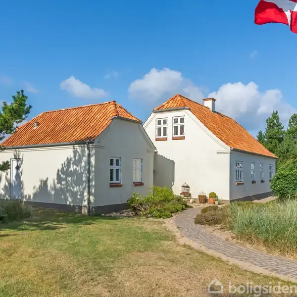Et traditionelt hvidt hus med rødt tegltag står i en frodig have med træer. En dansk flagstang til højre vejrer flaget under en klar blå himmel.