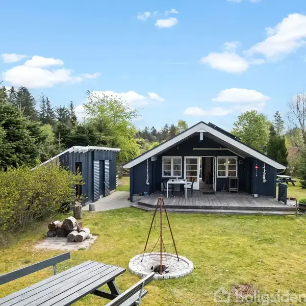 Et sort sommerhus med terrasse står i en grøn have omgivet af træer. Foran huset er der et bålsted og en bænk. Himlen er klar med spredte skyer.