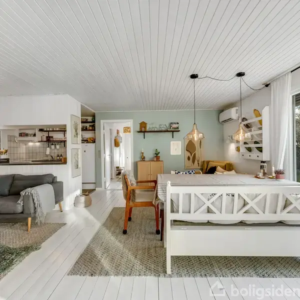 Stue med grå sofa til venstre, spisebord og bænke i midten. Rummets lyse træloft og gulv skaber en moderne atmosfære. Stor glasdør til højre åbner ud mod en have.