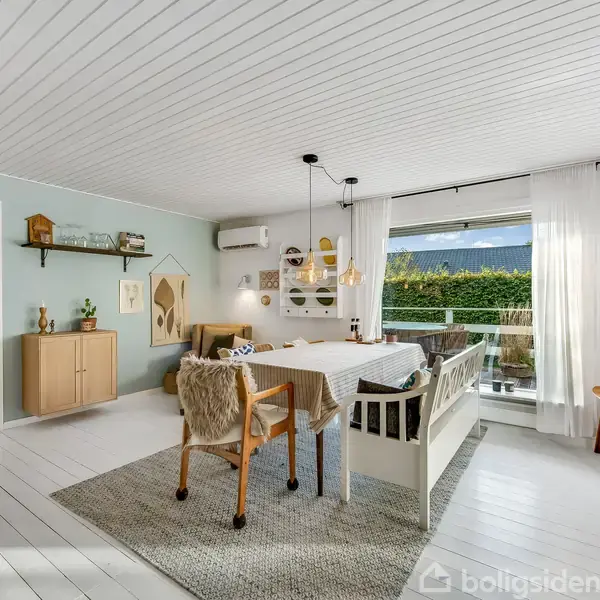 Spisebordet med stole omkring står i et lyst, stilfuldt indrettet rum med hvide vægge og gulve. Store vinduer med gardiner lader naturligt lys komme ind, mens planter og lamper skaber hygge.