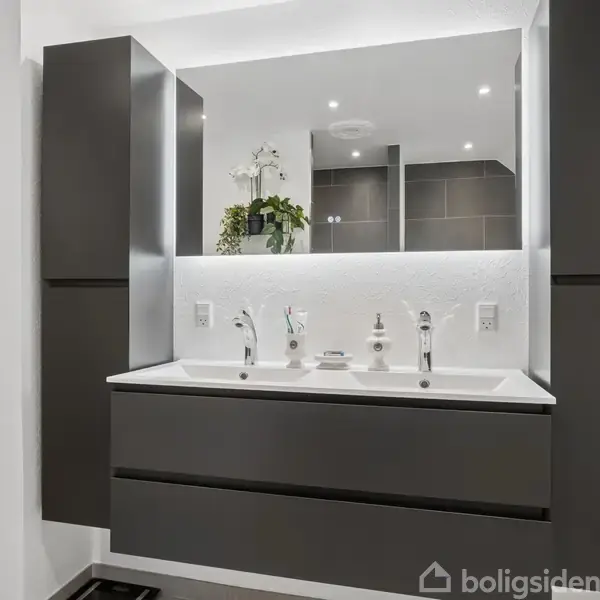 Et moderne badeværelse har en dobbel vask med grå underskabe, to spejle og planter ovenpå. Indbyggede spots belyser området, mens et beige håndklæde hænger til venstre.