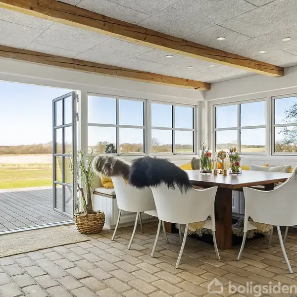 Spisebord med stole, belagt med lammeskind, står i en lys spisestue med store vinduer og åbne dobbeltdøre mod en træterrasse. Udsigt over en grøn mark.