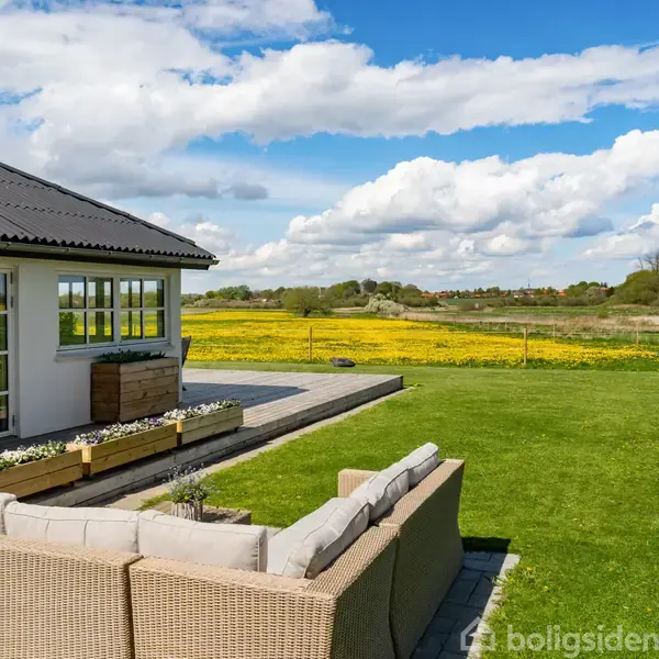 En sofa står på en terrasse ved et hus med store vinduer. Foran er en frodig græsplæne med udsigt over marker og træer under en blå himmel med hvide skyer.