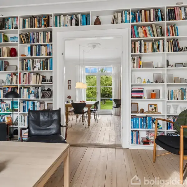 En bogreol fyldt med bøger og dekorative genstande står omkring en døråbning. Gennem døren ses en spisestue med spisebord og stole og en stor glasdør ud til en grøn have.