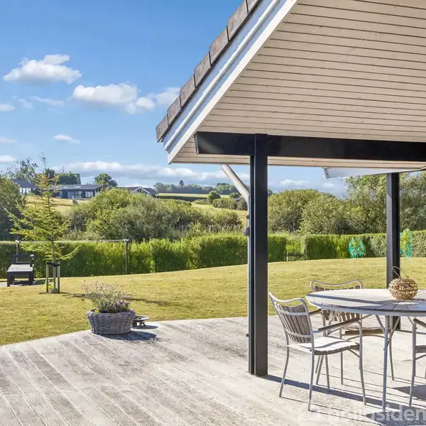Terrasse med spisebord og stole under overdækning. Græsplæne med haveudstyr i baggrunden. Omkranset af buske og træer i et fredeligt, solrigt landskab.