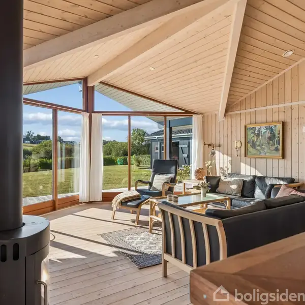 Træbrændeovn i hjørne brænder i en lys, moderne stue med højt til loftet, store vinduer, sort sofa og glasbord. Udsigt til grøn have og blå himmel.
