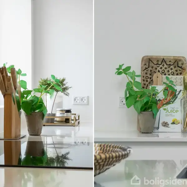 En køkkenbordsplade med planter i potter og træ-redskaber i glasbeholdere. En bog med titlen "Juice" står ved siden af en dekorativ bakke. Hvid og minimalistisk indretning.