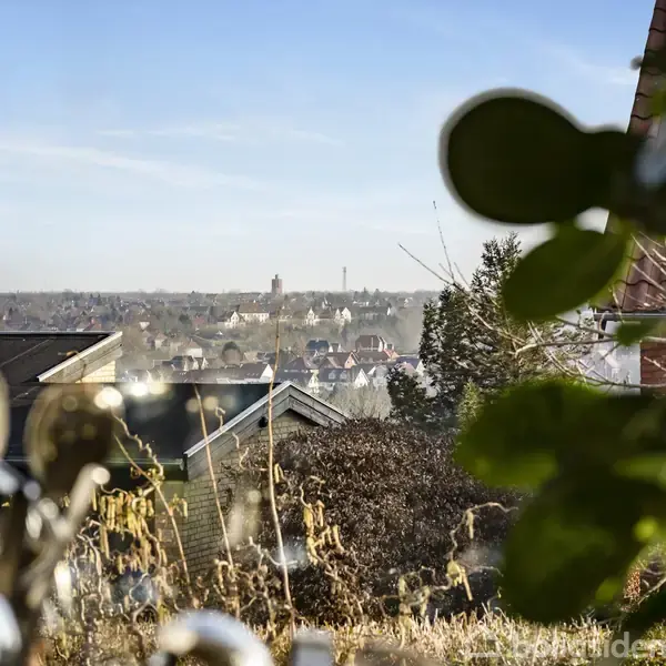 Vinduesramme indrammer en udsigt over en by med hustage og en fjern kirke i horisonten, omgivet af træer og planter tæt på vinduet.