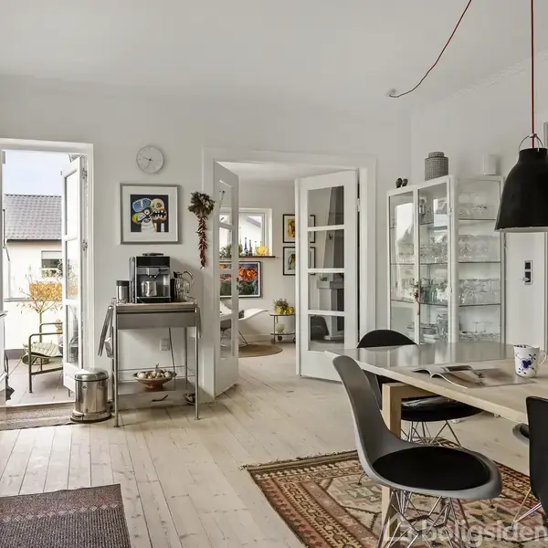 Køkkenbord med sorte stole står i et lyst rum med lyse trægulve. Kaffemaskine er placeret på et metalbord, og døre tillader udsigt til en tilstødende stue.