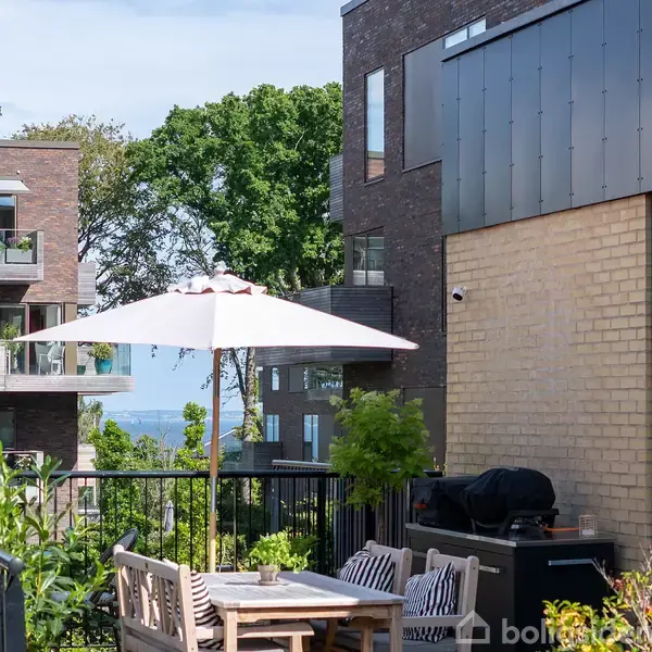 Terrassebord og stole under parasol står stille på altan omgivet af bygninger og grønne planter. Solskin kaster skygger, og udsigten viser træer og en anelse af himlen i baggrunden.