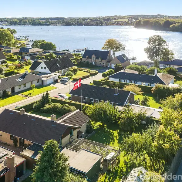 Et parcelhuskvarter ligger ved en sø, med huse, træer og haver. En dansk flagstang markerer en af haverne. Landskabet er frodigt og solbeskinnet med udsigt over vandet.