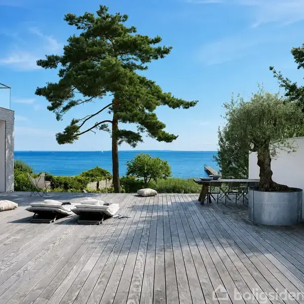 To liggestole står på en træterrasse med puder og et lille bord i nærheden. I baggrunden ses havet og træer, hvilket skaber en fredfyldt atmosfære.