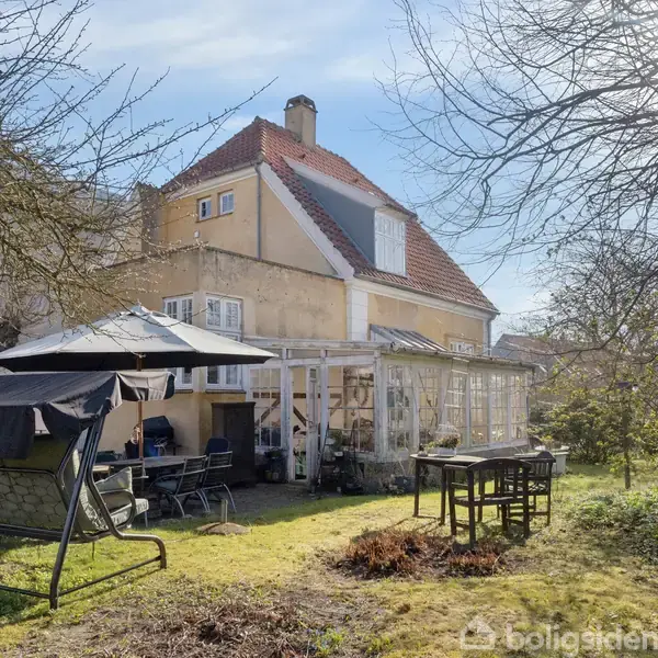 Hus med rødt tag dominerer billedet, omgivet af en have med flere træer og buske. En overdækket terrasse rummer havemøbler, hvoraf én er en havesofa under en parasol.