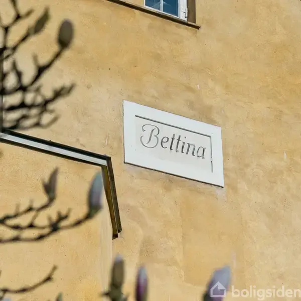 Bygning med gul facade, viser navnet "Bettina" på et skilt. Sollys belyser væggen, mens grene er i forgrunden. Vinduer ses øverst og til højre.