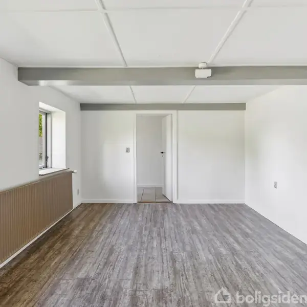 Rum med trægulv, hvidmalede vægge og to små vinduer. Midt i rummet ses en dør, og en synlig bjælke løber langs loftet.