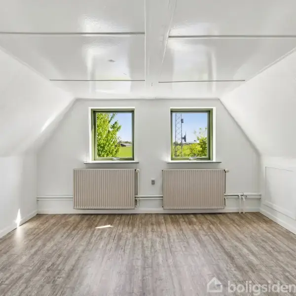 Et lyst, tomt loftsrum har trægulv og to vinduer, som giver udsigt til grønt landskab. Rummet er omgivet af hvide vægge og radiatorer under vinduerne.