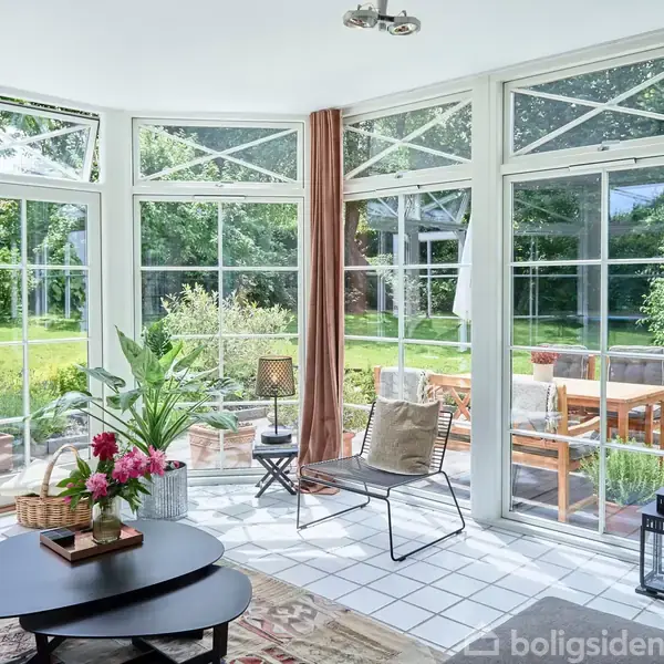 En lys udestue med store vinduer viser udsigt til en frodig have. Et bord med blomster, en stol og lanterner står på flisegulvet. Ingen tekst er synlig.