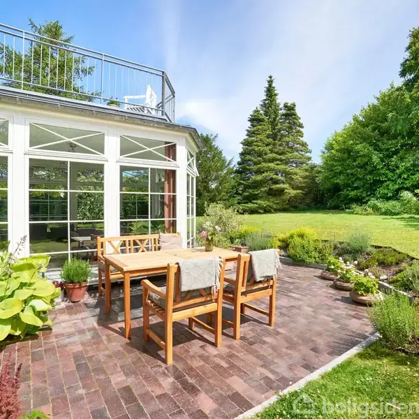 Et sæt træmøbler står på en flisebelagt terrasse foran et drivhus med glasvægge. Omgivelserne er en grøn have med græs og buske.