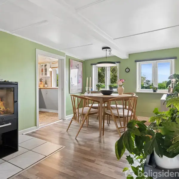 En spisebord samling står centralt, omgivet af grønne vægge og en vinduesudsigt. En brændeovn i hjørnet brænder. Planter og udsmykning skaber en hyggelig stue-atmosfære.