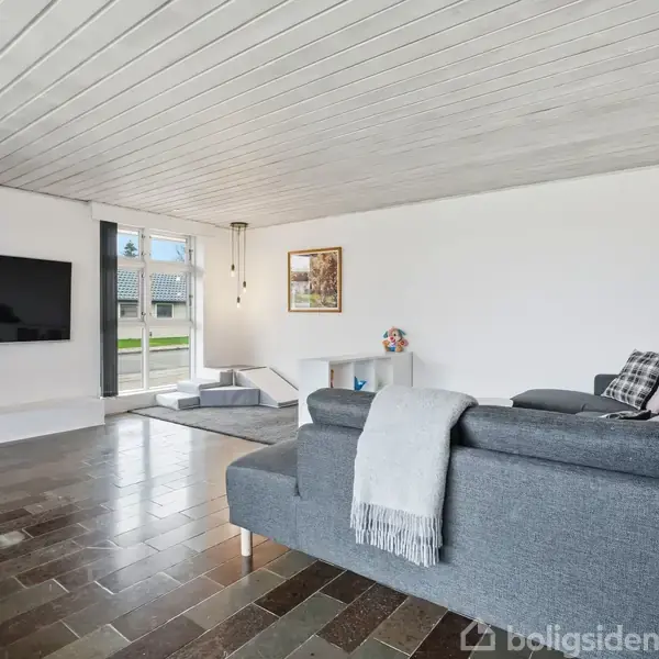 En moderne stue har en grå sofa med tæpper, der står mod en fladskærms-tv på en hvid væg. Rummet er lyst med store vinduer og minimalistisk indretning.